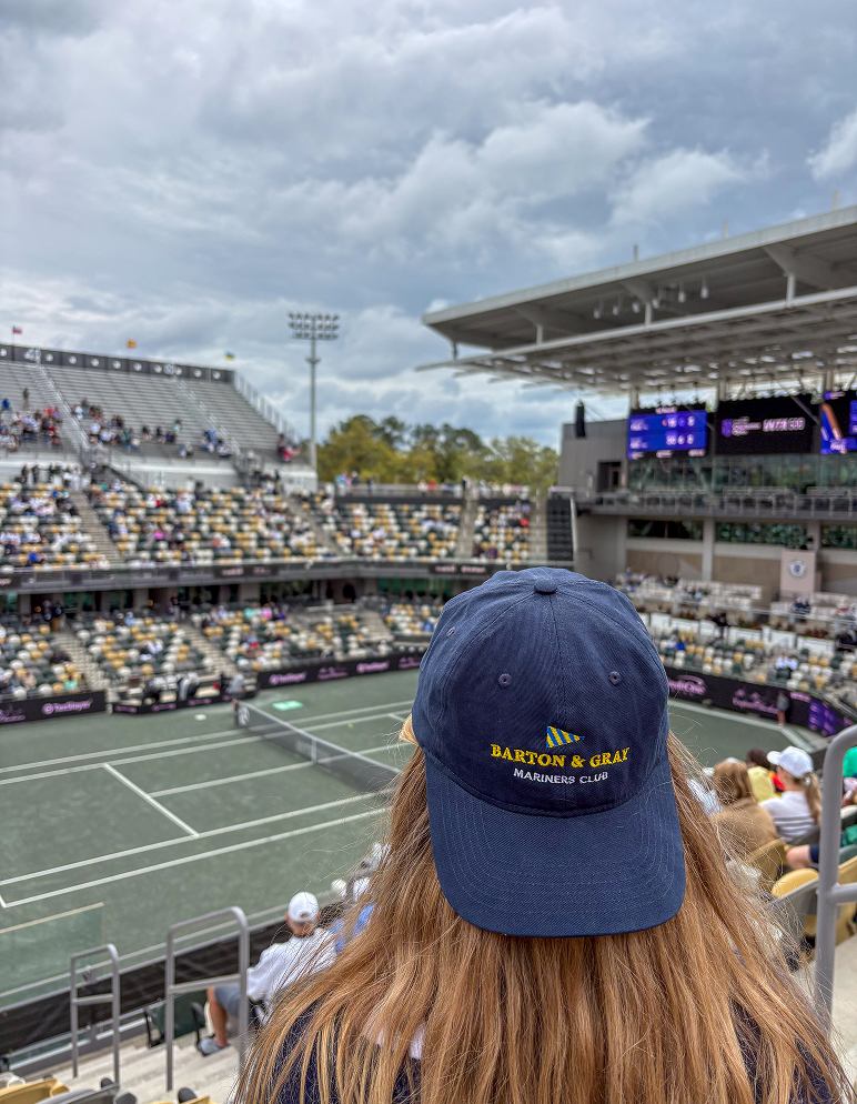 Woman wearing Barton & Gray Mariners Club navy hat watching professional tennis match from stadium seats in Charleston, SC