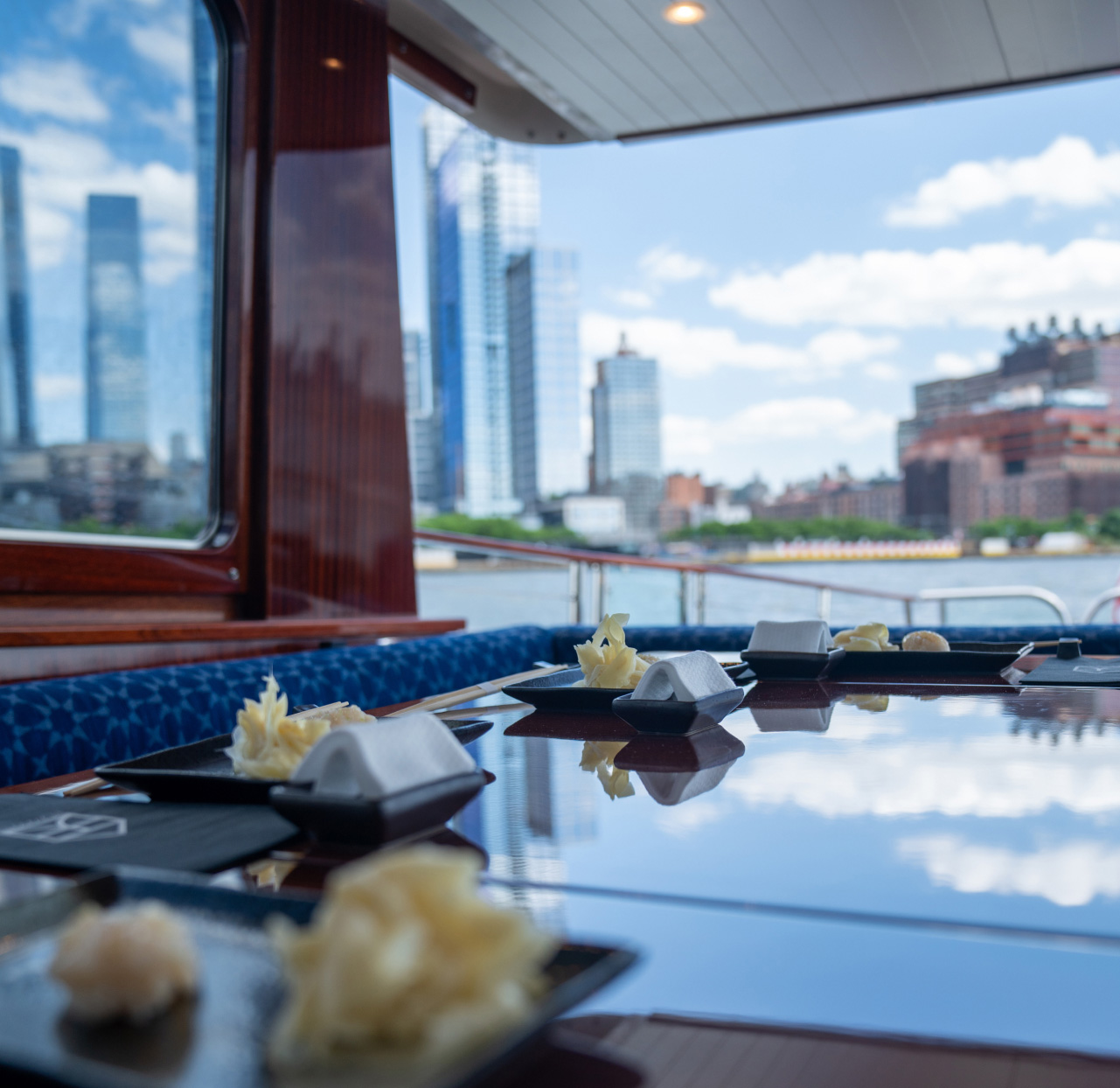 Chef's omakase course place settings on a Barton & Gray yacht deck with a reflective table and a city skyline across the water – luxury onboard sushi dining